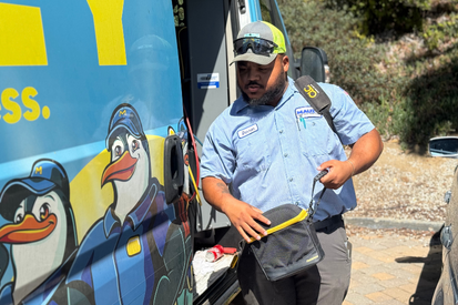 A worker in uniform stands beside a van with cartoon penguin graphics, holding a shoulder bag and wearing sunglasses, a cap, and a name badge.